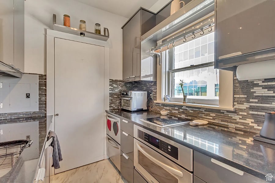 Sleek, sun-filled galley kitchen featuring quartz countertops, high-gloss gray cabinetry, a glass-tile backsplash, and modern stainless-steel appliances.