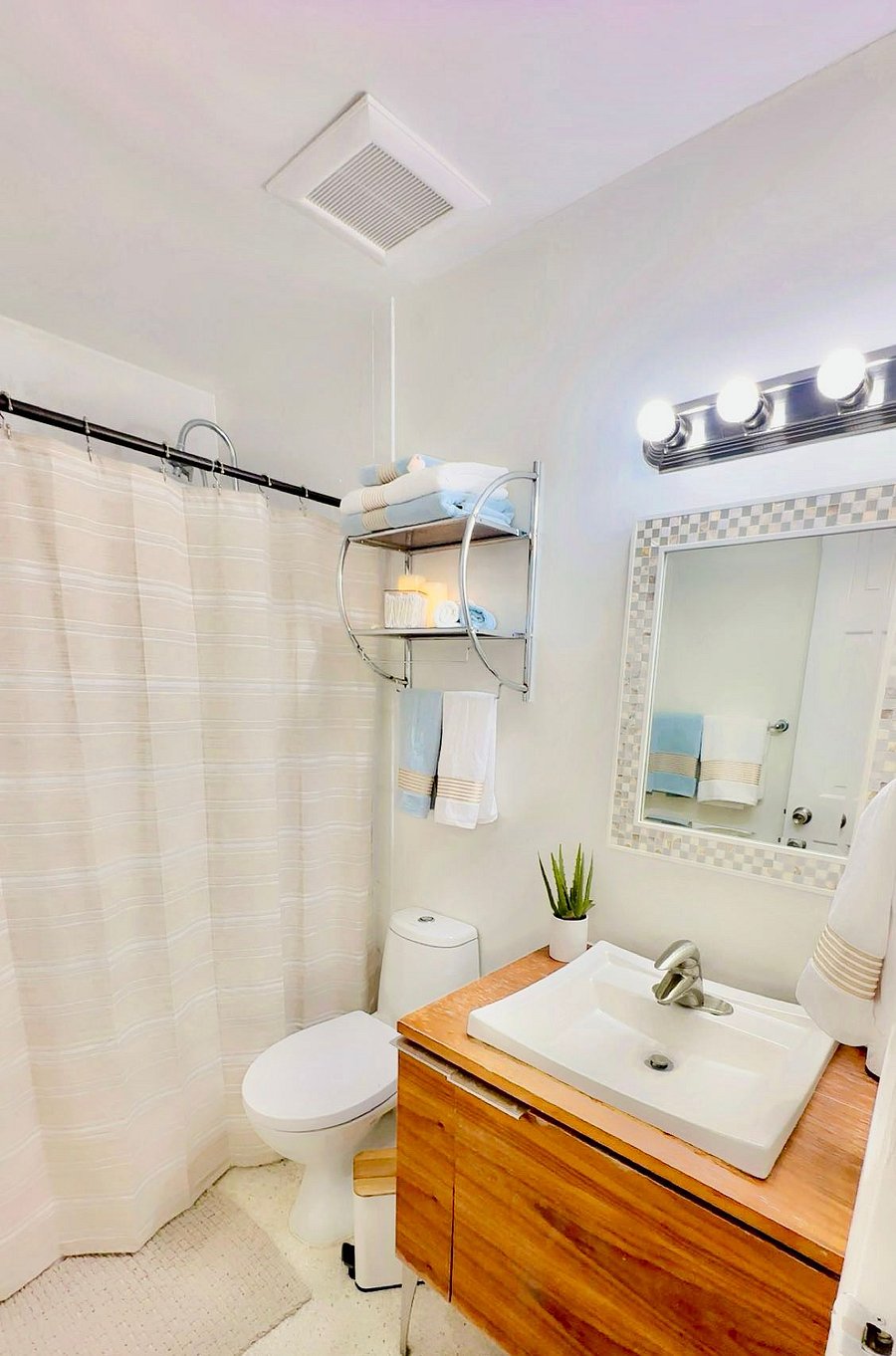 The second bathroom includes a vanity, shower, and overhead lighting with a framed mirror. A shelf holds towels and supplies. Durable terrazzo flooring keeps the space cool and easy to clean. A hallway just outside features a full-length wall mirror and soft runners, creating a functional and private transition to the second bedroom.