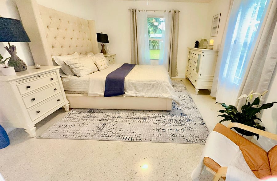 Second bedroom with a queen bed, blackout curtains, ceiling fan, dresser, and two windows one with a view of the backyard. Includes a lounge chair, mirrored closet, and leafy plant. Restored Florida terrazzo floors keep it functional and clean. Quiet and ready for extended stays.