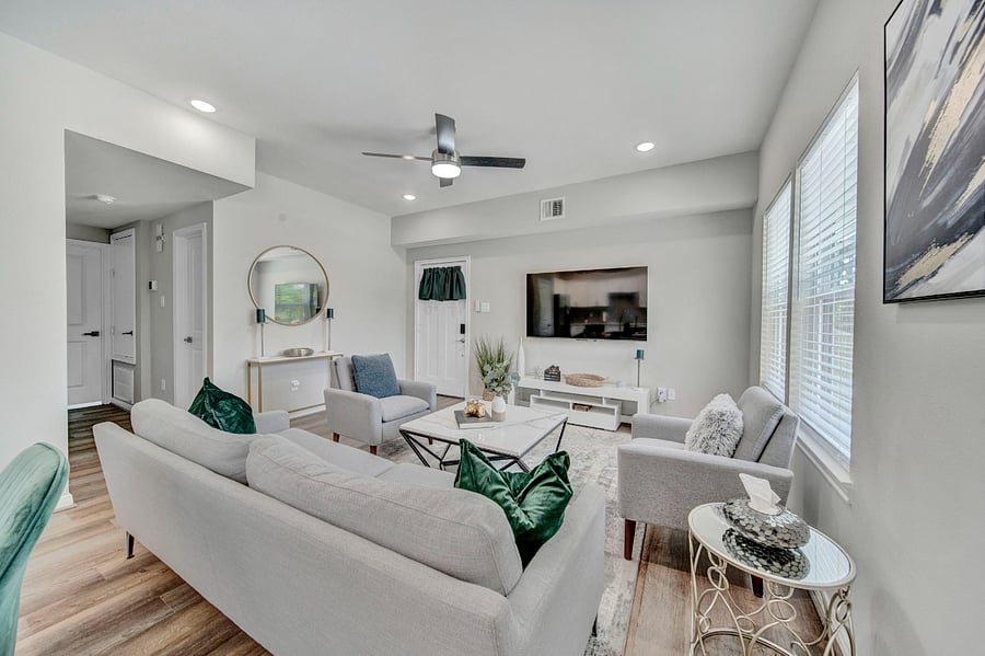 The seating area includes a white sofa set adorned with green cushions, complemented by a coffee table in the center and two armchairs. Opposite the seating area, a 75" flat-screen TV is mounted on the wall, providing high-definition entertainment.