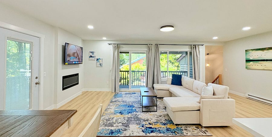 Bright open-concept living room with a comfortable sectional sofa, seamlessly connected to a modern kitchen and dining area, all framed by large windows highlighting lush surrounding trees