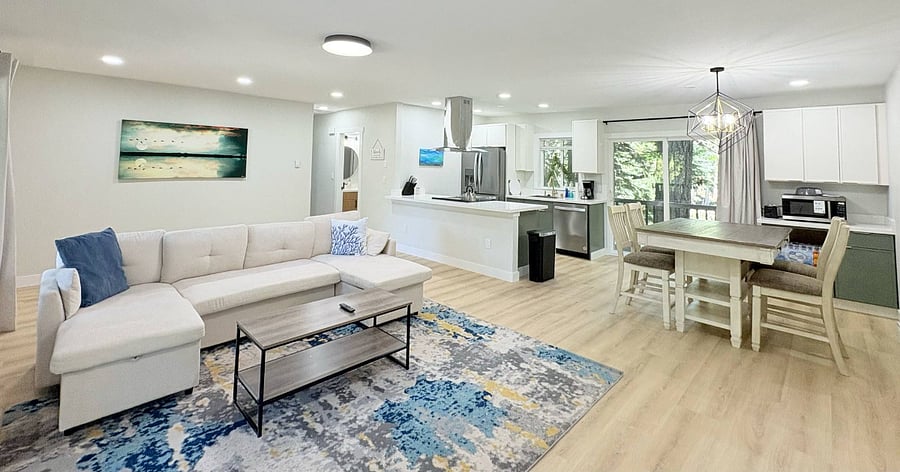 Bright open-concept living room with a comfortable sectional sofa, seamlessly connected to a modern kitchen and dining area, all framed by large windows highlighting lush surrounding trees