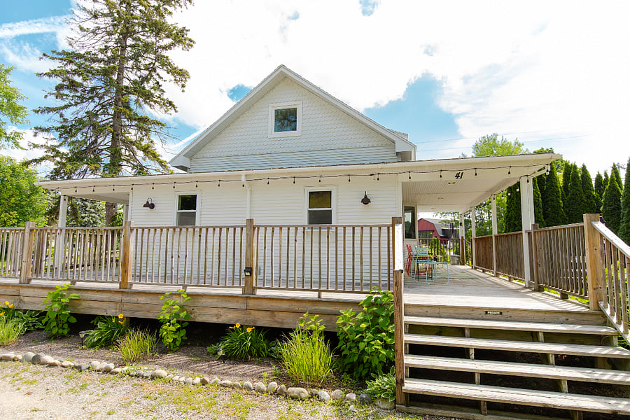 The west-facing side of the cottage with stairs up to the porch from the parking area. Bistro lights line the wraparound porch for a cozy atmosphere, whether being enjoyed from inside or outside the house.