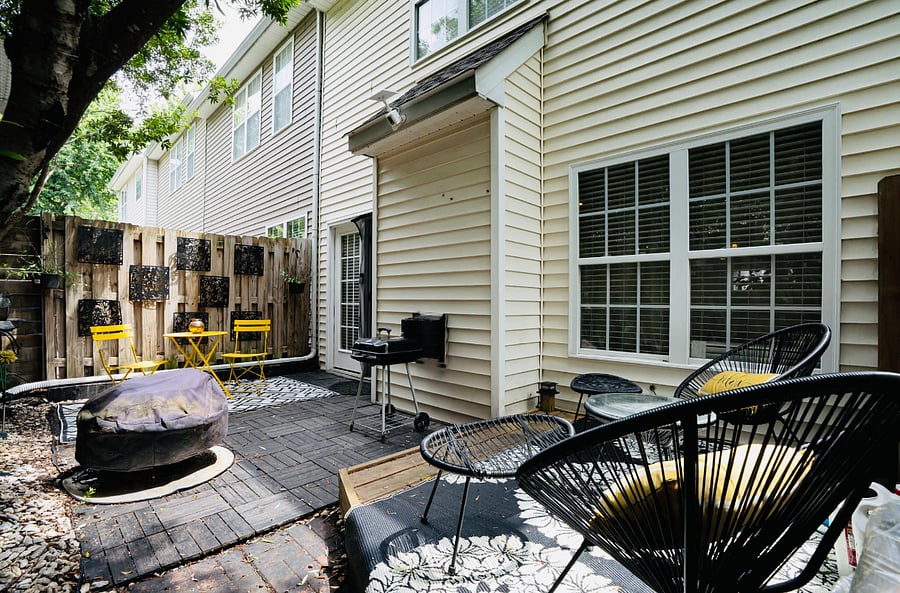 Unwind in this cozy, secluded patio oasis tucked away in a serene corner. The space is framed by lush trees that lend a natural, calming ambience. A compact, rustic wood deck anchors the area, with comfortable seating with cushioned wicker chairs.