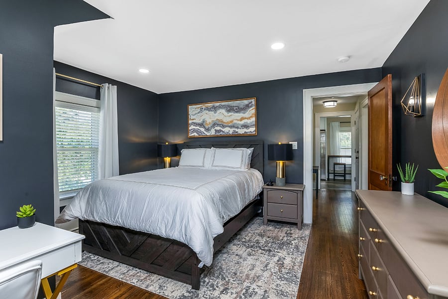 The large bedroom features a queen bed, two nightstands with lamps, a large dresser with mirror, and a work area that includes a desk, chair, and another cushioned window seat