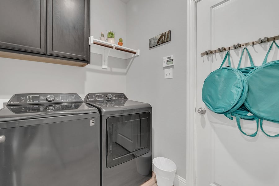 Laundry room with modern high-capacity washer and dryer, overhead cabinetry, and wall hooks for organized storage. Laundry hampers, iron, and ironing board. Initial supply of laundry detergent and dryer sheets.