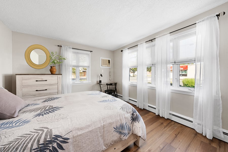 Bright front queen bedroom featuring a dedicated workstation, wall air conditioner, windows, and room-darkening shades.
