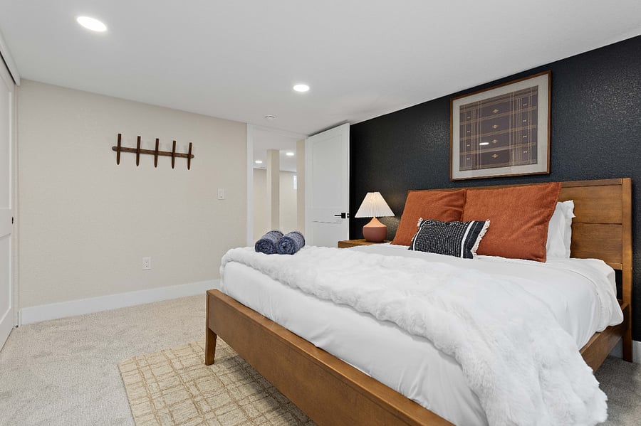 This large bedroom features a warm wood bed frame, soft layered bedding, and a dramatic dark navy accent wall for contrast. The modern lighting and simple decor create a peaceful retreat.