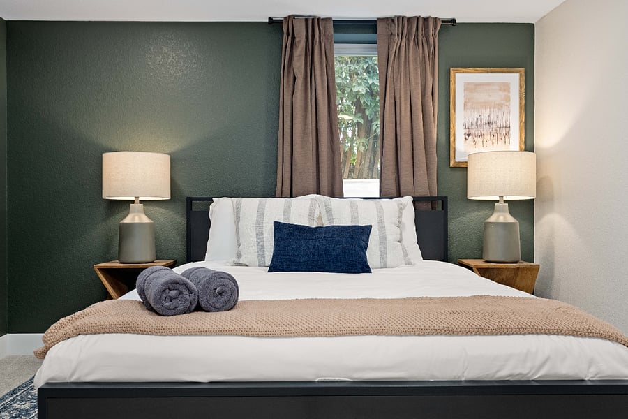Stylish and serene bedroom featuring a black bed frame, neutral bedding with a navy accent pillow, and matching wood nightstands with modern lamps. The rich green accent wall adds a grounded, natural touch to the space.