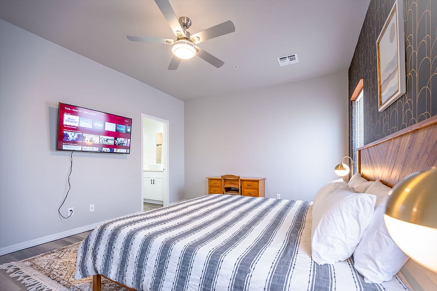 View of primary suite from right side of the room showing the door next to TV that leads to bathroom and walk-in closet