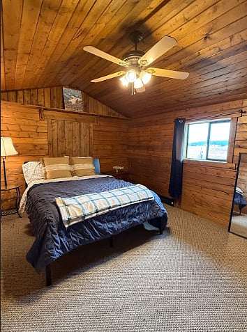 Bedroom with queen bed, bedside table with lamp. Dresser. Headboard has plug outlets. Electric baseboard heating for winter and ceiling fan for summer. Bed is lifted to include storage space.