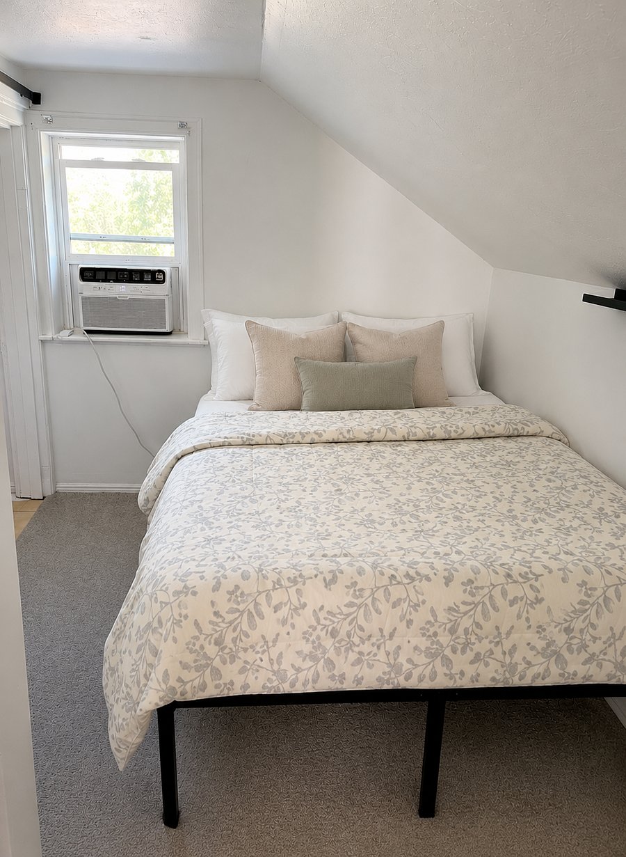 Queen bed in a cozy, bright bedroom with soft linens and blackout shades for added comfort.