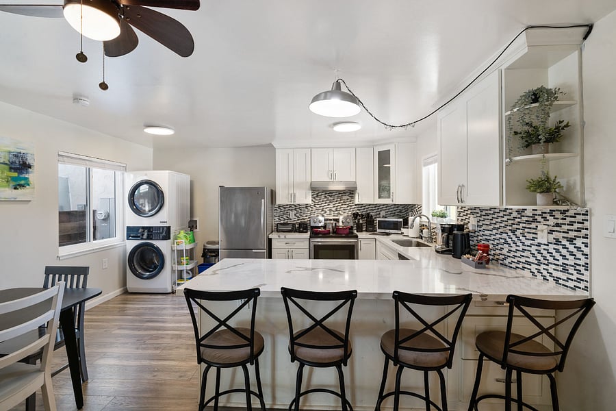 Modern kitchen with white cabinets, stainless steel appliances, stylish backsplash, center island with bar stools, ceiling fan, and washer/dryer in the corner.