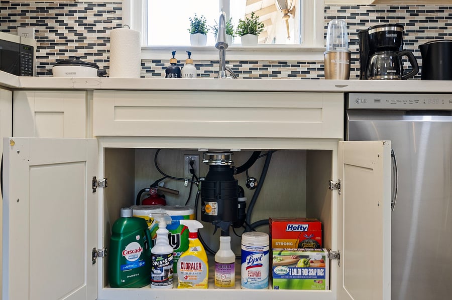 A cabinet under the sink reveals cleaning supplies like Cascade detergent, Cloralen bleach spray, Lysol wipes, and Hefty trash bags. The garbage disposal unit and plumbing are visible inside the cabinet, with a fire extinguisher in the back.
