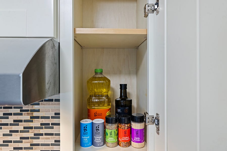 An open kitchen cabinet with cooking essentials: a bottle of vegetable oil, a dark bottle of balsamic vinegar or soy sauce, and six spice containers including iodized salt, ground black pepper, ground red pepper, and garlic powder.