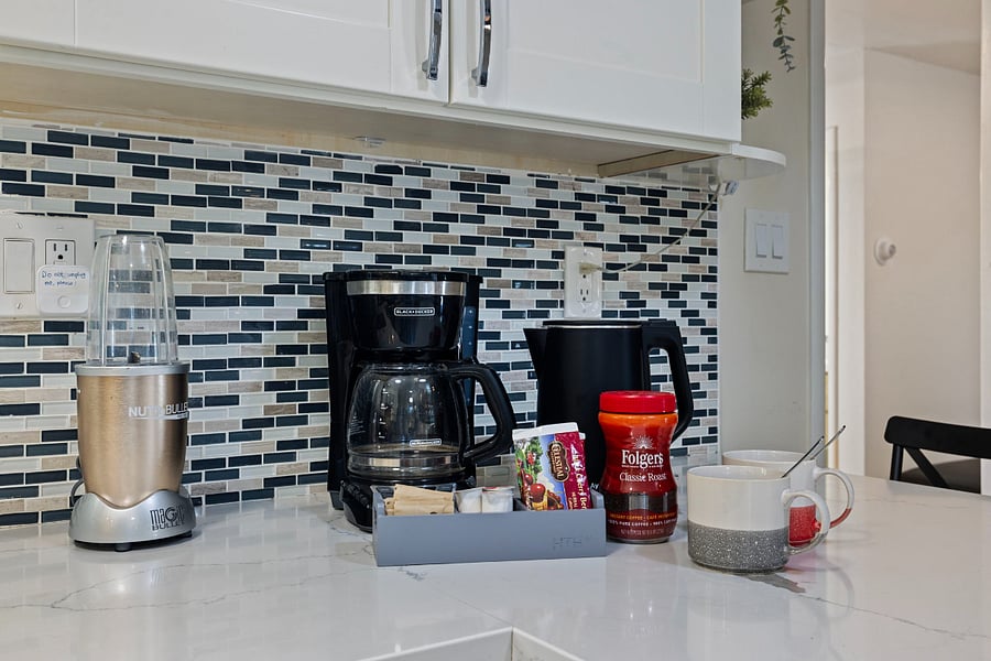 A kitchen counter with a NutriBullet blender, a black Hamilton Beach coffee maker, a black electric kettle, Folgers Classic Roast coffee, and two mugs.