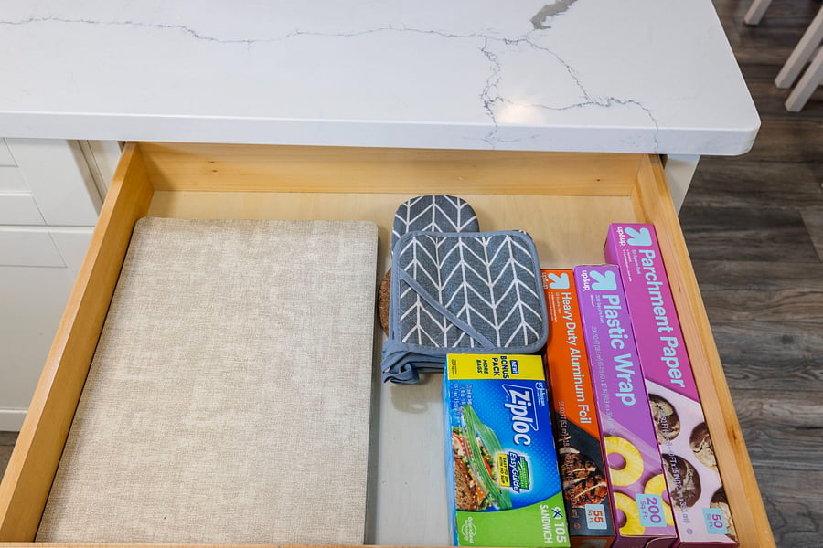 An open kitchen drawer containing a beige cutting board, gray patterned oven mitt and pot holder, and boxes of parchment paper, plastic wrap, heavy-duty aluminum foil, and Ziploc sandwich bags, all neatly organized under a white marble countertop.