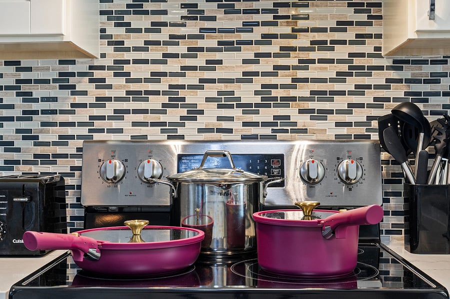 A modern kitchen stove with a stainless steel backsplash and black, white, and beige tile wall. Two purple pots with glass lids and gold handles sit on the stovetop. Nearby are a black toaster and a container holding cooking utensils.