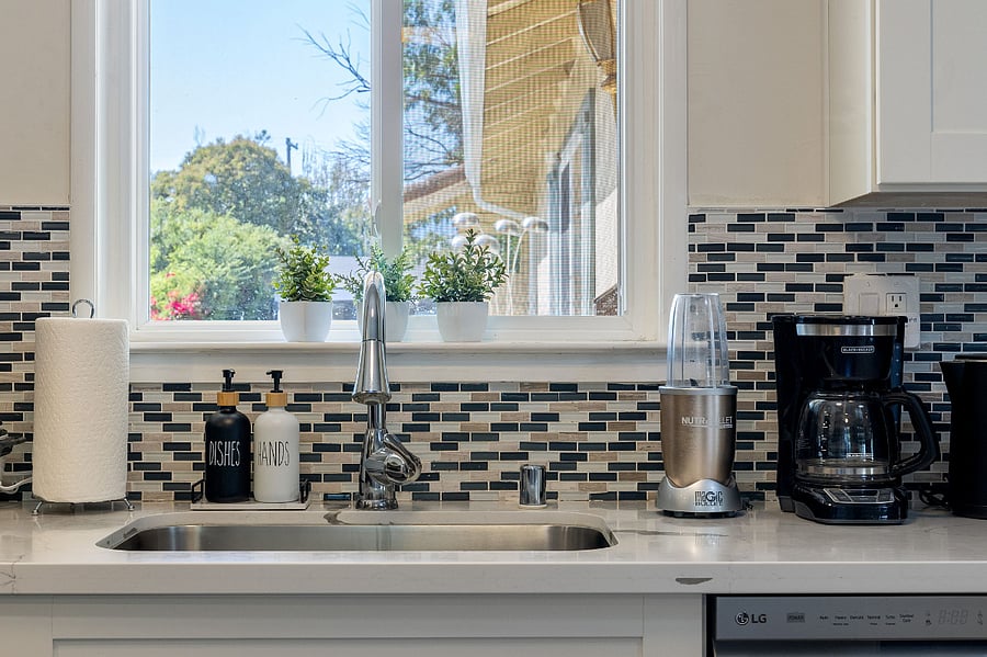 A clean kitchen sink area with a stainless steel faucet and under-mount sink. The countertop holds a paper towel roll, soap dispensers, a blender, and a coffee maker.