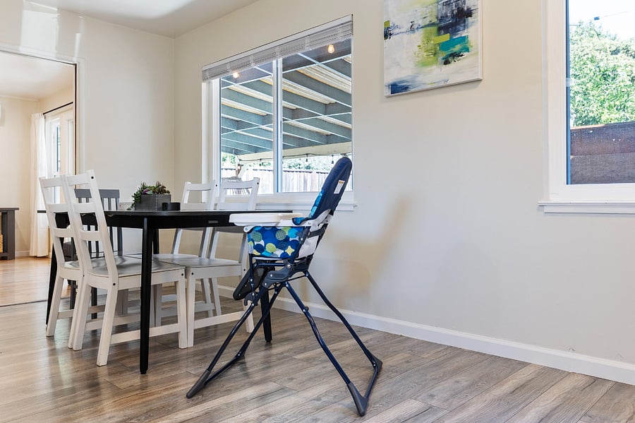 A black dining table with white and gray chairs sits near a window. A colorful high chair is positioned at the table.