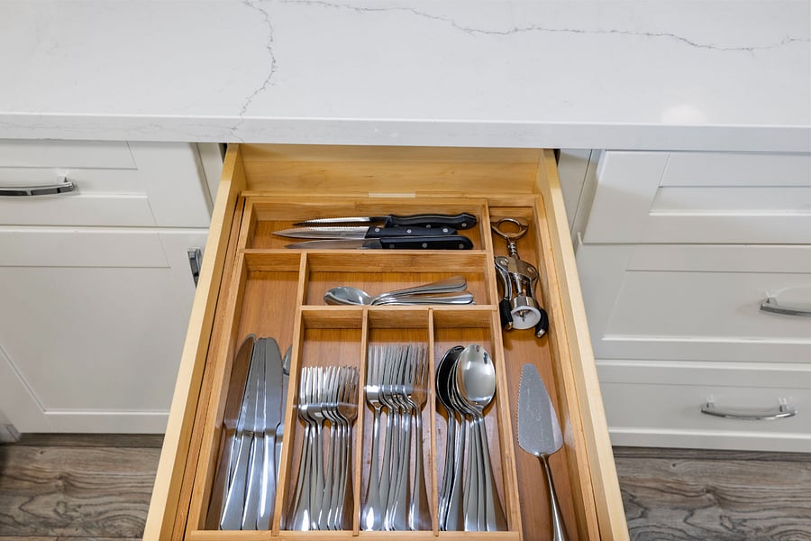 An open wooden kitchen drawer with neatly organized stainless steel cutlery: knives, forks, spoons, teaspoons, a cake server, three kitchen knives, and a corkscrew, all arranged in separate compartments under a white marble countertop