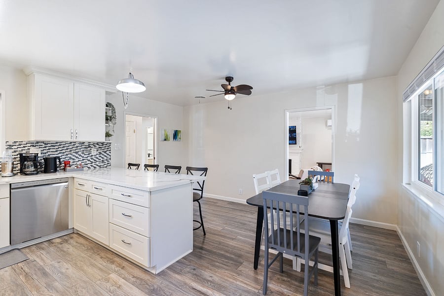 Bright modern open kitchen and dining area with white cabinetry, large island with drawers, mixed white and gray chairs, and wood-style laminate flooring.