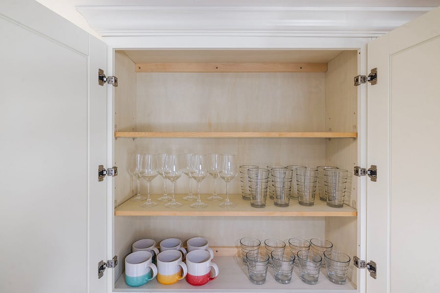A white kitchen cabinet with open doors showing three shelves. The middle shelf holds six wine glasses and eight striped drinking glasses. The bottom shelf has six colorful mugs and eight short drinking glasses, all neatly arranged inside