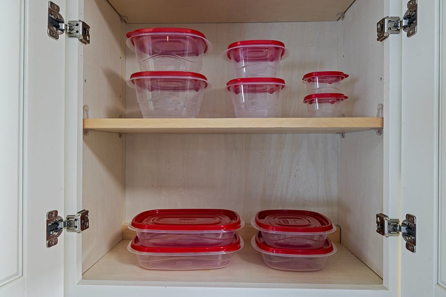 A white kitchen cabinet with two shelves holds various clear plastic food storage containers with red lids. The containers are neatly stacked by size, with larger containers on the bottom shelf and smaller ones on the top shelf.
