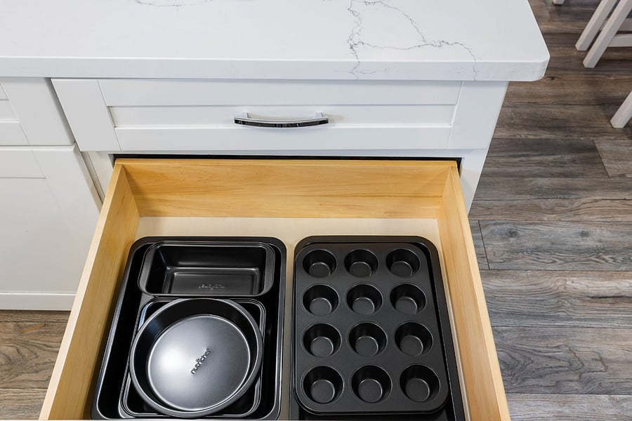 A wooden kitchen drawer beneath a white marble countertop contains various black baking pans, including a loaf pan, round cake pans, and a 12-cup muffin tin, neatly organized on a wooden floor background.