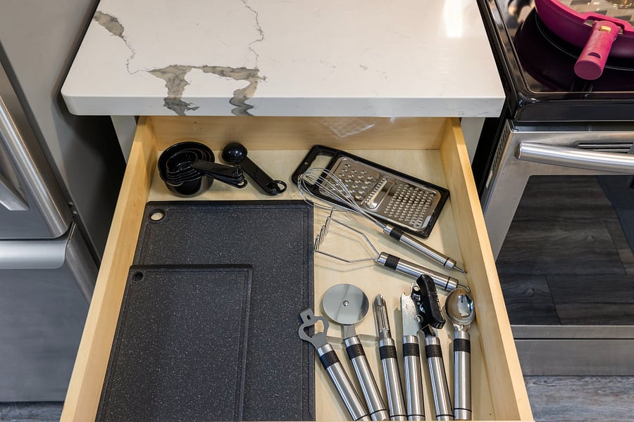 A wooden kitchen drawer under a white marble countertop holds black cutting boards, black measuring spoons, a metal grater, two whisks, and stainless steel utensils including a pizza cutter, can opener, vegetable peeler, and ice cream scoop.