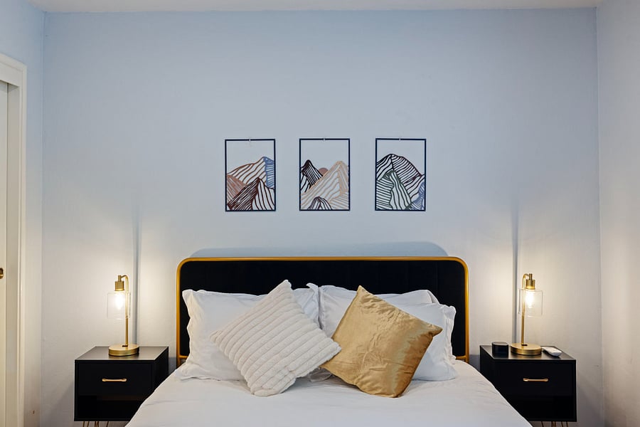 A stylish bedroom with a black and gold bedframe, white bedding, and gold and cream pillows. Two black nightstands with modern gold lamps flank the bed. Three framed abstract mountain artworks in black and earthy tones adorn the light blue wall.