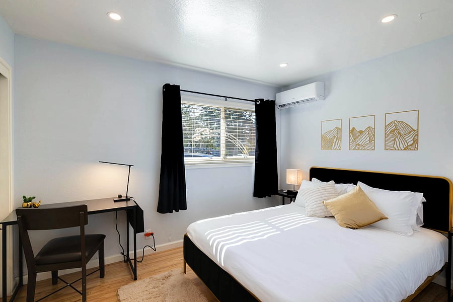 Bright bedroom with light blue walls, large bed with white bedding and beige pillows, black desk and chair by the window with black curtains, and three gold-framed abstract art pieces above the bed.
