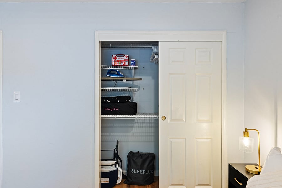 A small closet with sliding white doors, one open to reveal organized shelves. Items include a first aid kit, an iron, a black bag, and a "SLEEPLUX" storage bag