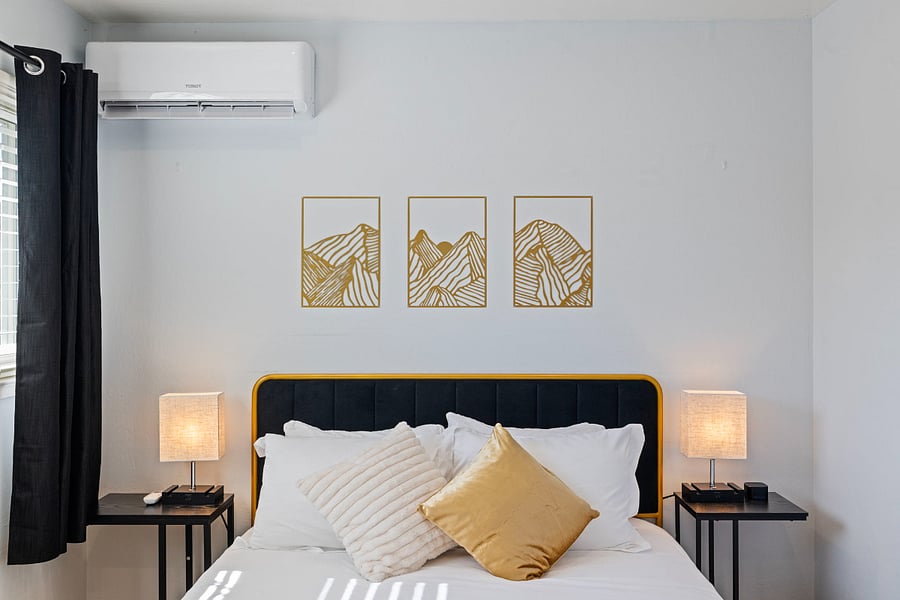 A cozy bedroom with a black and gold headboard bed, adorned with white and gold pillows. Two bedside tables hold lamps. Above the bed, three gold-framed mountain line art pieces decorate the white wall. Black curtains frame the window.