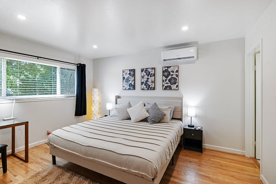 Master bedroom with a large bed dressed in striped bedding and multiple pillows. Two nightstands with lamps flank the bed. Three floral artworks hang above the headboard. A window with black curtains, and a small desk complete the room.
