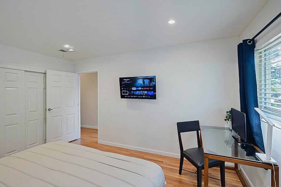 A master bedroom with a striped bedspread, a wall-mounted TV, and a glass desk with a black chair. A window with navy curtains.