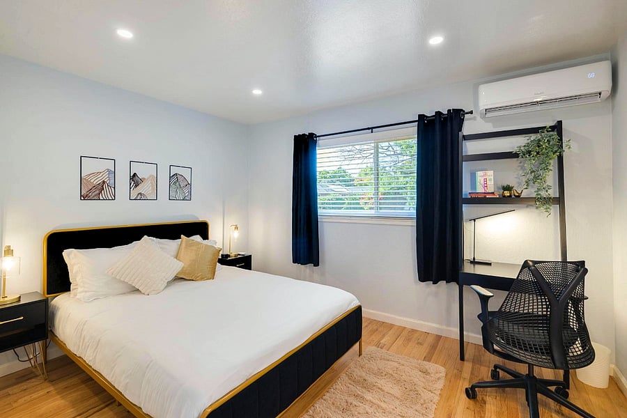 A bright bedroom with a black and gold bed, white bedding, and gold and cream pillows. Black nightstands with gold lamps flank the bed. A window with black curtains. A black desk and chair sit under an air conditioner