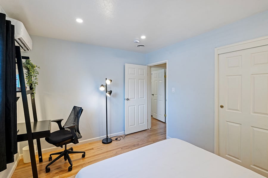 A light blue bedroom corner with a white door and closet. A black desk and mesh office chair sit near a window with black curtains. A three-bulb floor lamp stands beside the door. The room has wood flooring and recessed ceiling lights.