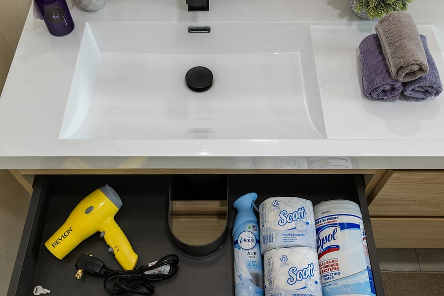A bathroom drawer beneath a white sink holds a yellow Revlon hair dryer, a blue Febreze air freshener, two rolls of Scott toilet paper, and a container of Lysol disinfecting wipes.