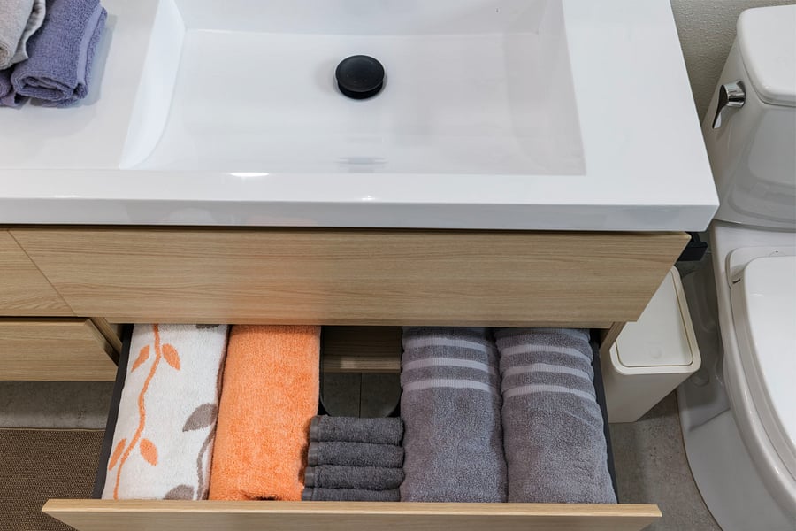 A bathroom drawer neatly organized with folded towels: two large gray towels, several small gray washcloths, an orange towel, and a white towel with an orange leaf pattern. The drawer is under a white sink with a wooden cabinet.