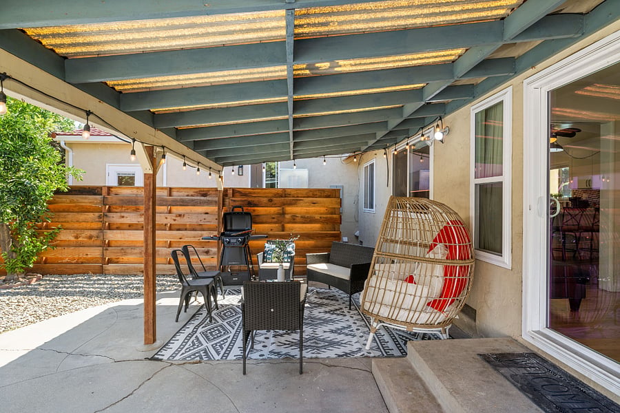 Covered patio with teal beams, hanging egg chair with red cushions, black wicker chairs, small table, grill, and stylish patterned rug. Sliding glass doors lead inside, with a wooden fence enclosing the space.