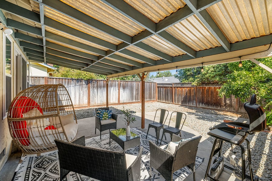 Covered patio with cozy wicker chairs, black loveseat, hanging egg chair with red cushions, patterned rug, and a grillperfect for relaxing and enjoying the fenced backyard with trees.