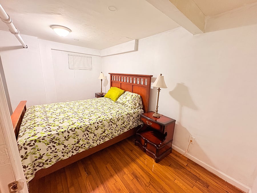 Cozy and comfortable furnished bedroom featuring a beautiful wooden queen size bed frame, matching nightstands,and warm hardwood flooring. The room offers a clean, inviting atmosphere with ample space around the bed and convenient lighting on both sides.