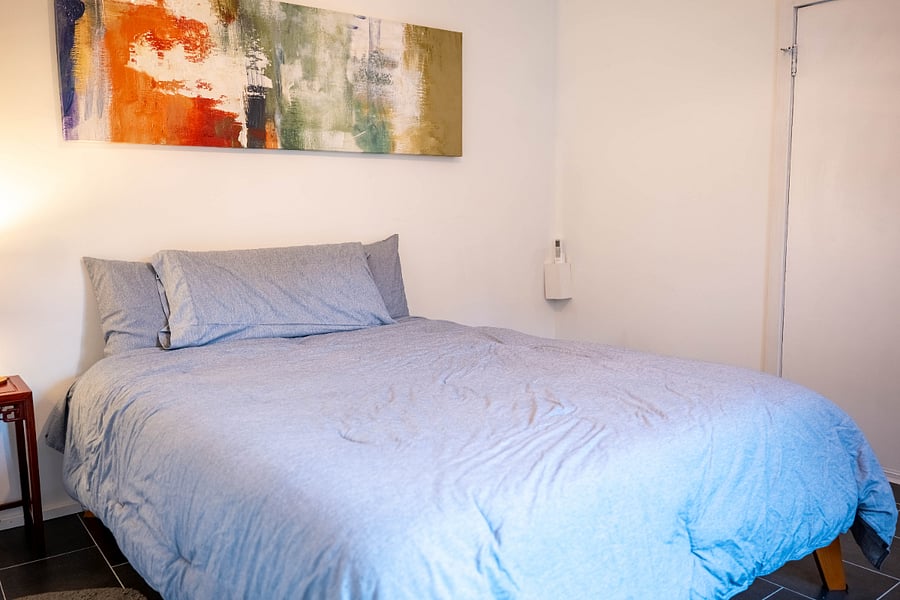 Third bedroom, queen memory foam mattress, personal airconditioner, exit to back yard, table lamp, bedstand table, rocking chair recliner, artwork