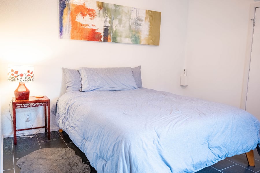 Third bedroom, queen memory foam mattress, personal airconditioner, exit to back yard, table lamp, bedstand table, rocking chair recliner, artwork