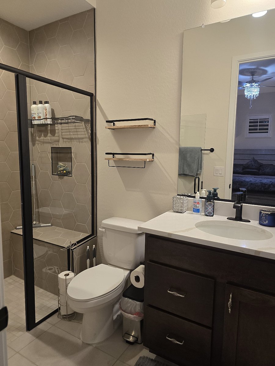 Room A Private Bathroom
This modern en-suite bathroom features a glass-enclosed shower with striking hex tile, a sleek vanity with white countertop, and a large mirror with bright overhead lighting. Thoughtful storage includes open shelving above the toilet and under-sink cabinetry. Personal care items are neatly organized in woven baskets, and guests have access to essentials like soap, cotton swabs, and a hair dryer. A decorative towel and candle add a warm, welcoming touch.