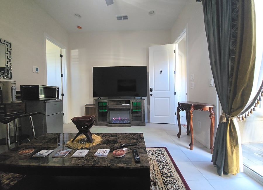Room A Private Lounge Area
This guest suite features a cozy lounge with a mounted flat-screen TV set directly above an electric fireplace, perfect for relaxing nights in. A marble-top coffee table anchors the space, surrounded by a leather sofa and warm accent lighting. The entertainment stand includes ambient green lighting and storage. A kitchenette with microwave and coffee maker adds everyday convenience. Sliding glass doors open to fresh air and natural light, with elegant curtains for privacy. Thoughtful touches like a decorative bowl, coasters, and a side table make the space feel complete.