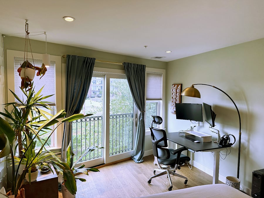 Standing desk with dual mounted monitors and an X-chair office chair.  Sun-soaked area to work next to a juliet balcony and large windows + plant corner.