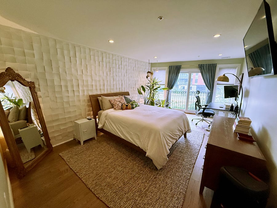 Master bedroom has a queen size sleigh bed with linen bedding and a cozy oversized down comforter. The bedroom accent wall has a mosaic tile pattern and backlights that alight the wall at nighttime.  Mounted smart tv + desk area and plant corner with a beautiful soft-colored grow light.