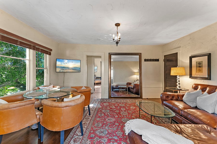 View from living room looking into bedroom: Smart TV, wood blinds, oversized floor mirror, comfy leather love seat sectional with cushy pillows and soft cable knit blanket. Two side tables with lamps. Glass top coffee table. Glass top dining table with 4 chairs.